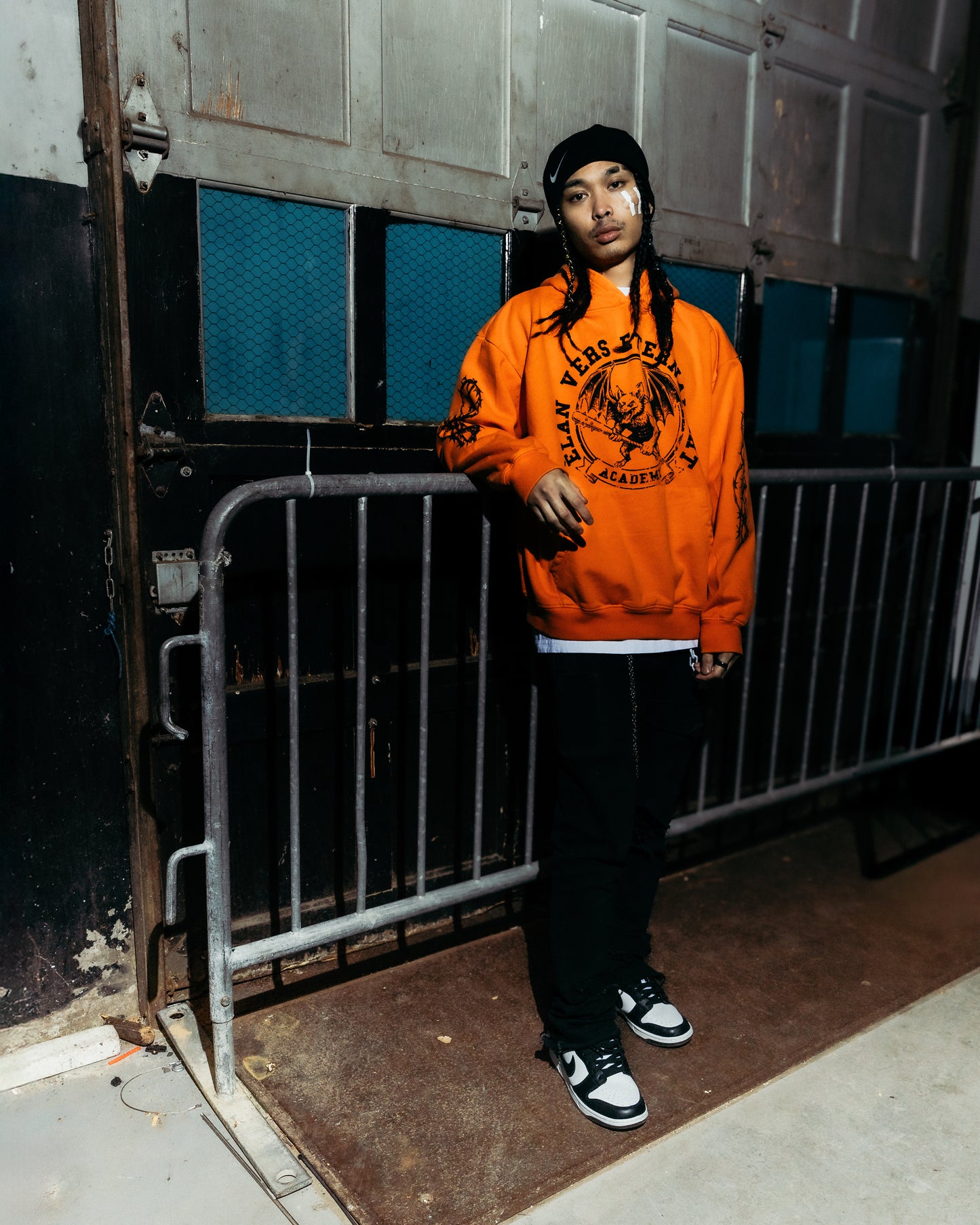 Asian model wearing an orange sweatshirt with a graphic design, standing in front of a metal gate.