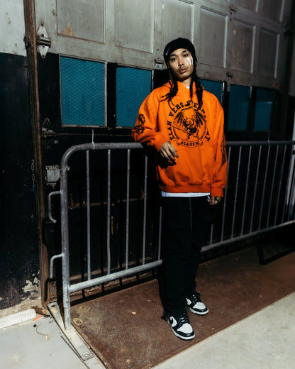 Asian model wearing an orange sweatshirt with a graphic design, standing in front of a metal gate.
