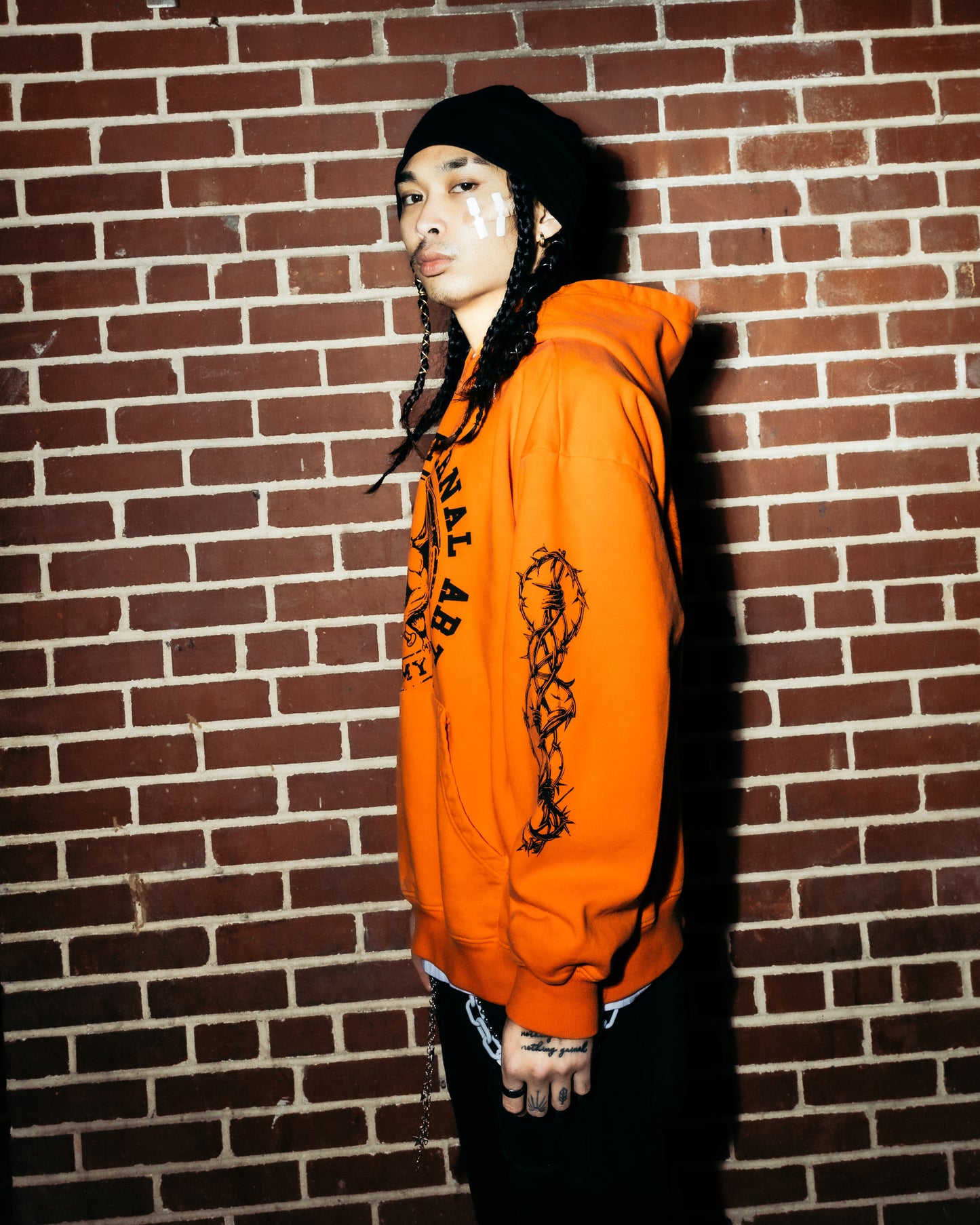 Asian model wearing an orange hoodie with a graphic design, standing against a brick wall.