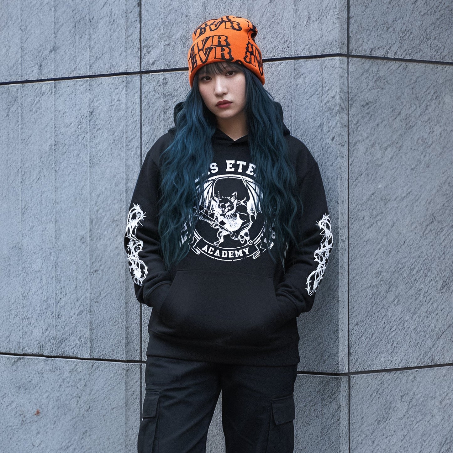 Woman wearing a black hoodie with white graphics and text, black pants, and black boots against a gray wall.