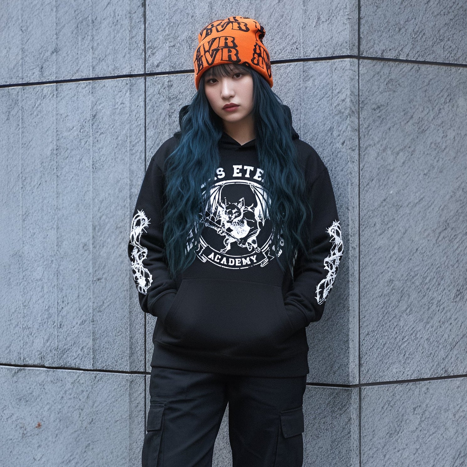 Woman wearing a black hoodie with white graphics and text, black pants, and black boots against a gray wall.